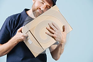 Cheerful delivery man in uniform is hugging cardboard box with his eyes closed photo