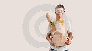 Cheerful disabled boy with Down syndrome smiling at camera while holding paper bag with food, standing isolated over