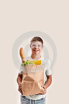 Cheerful disabled boy with Down syndrome smiling at camera while holding paper bag with food, standing isolated over