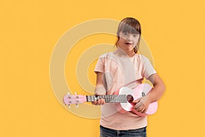 Cheerful disabled girl with Down syndrome looking at camera while playing ukulele, standing isolated over yellow