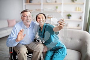 Cheerful disabled senior man in wheelchair and his nurse taking selfie indoors, selective focus