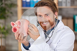 cheerful doctor holds stethoscope to piggy-bank
