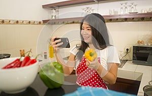 Cheerful domestic cooking - young happy and beautiful Asian Chinese home cook woman in red apron reading healthy recipe in mobile