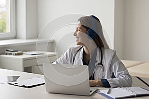 Cheerful dreamy pretty doctor in headphones working at laptop