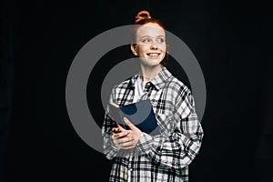 Cheerful dreamy young woman student holding opened book and looking away on black background.