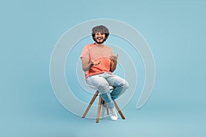 Cheerful eastern guy sitting on chair over blue background