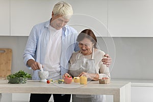 Cheerful elder husband helping mature senior wife to cook dinner