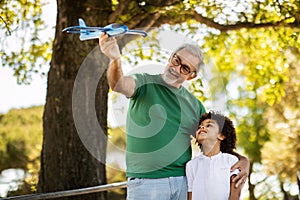 Cheerful elderly european grandfather and mixed race little boy play toy airplane in park, enjoy free time