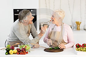 Cheerful Elderly Husband Feeding Wife Tasting Dinner Cooking In Kitchen