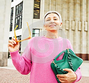 A cheerful elderly lady in a pink sweater is relaxing on the street on a summer day.