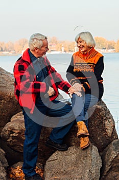 Cheerful elderly people a woman and a man are sitting on the stones and hugging on the lake, against the background of the bridge