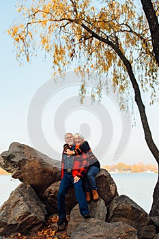 Cheerful elderly people a woman and a man are sitting on the stones and hugging on the lake, against the background of the bridge