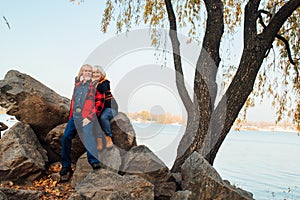 Cheerful elderly people a woman and a man are sitting on the stones and hugging on the lake, against the background of the bridge