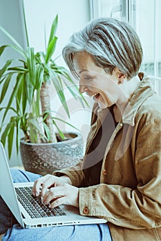 Cheerful elderly woman is chatting online, she laughs typing on laptop keyboard. Charming graying model sitting on
