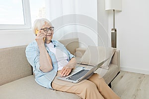 a cheerful elderly woman in glasses and a light shirt talking on the phone is working at a laptop enjoying the benefits