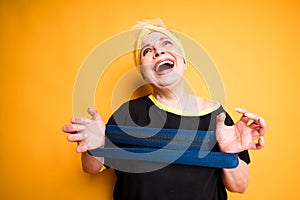 Cheerful elderly woman laughing and stretching exercise elastic band in hands while standing on yellow background