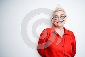 Cheerful elderly woman with mohawk wearing glasses and in a red shirt smiling with hands behind her back photo