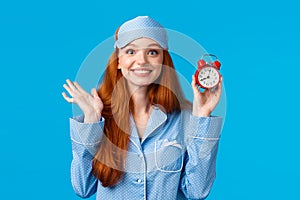 Cheerful and enthusiastic redhead girl dont want to oversleep, set alarm clock, holding red watch and smiling joyfully