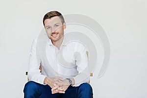 Cheerful entrepreneur, formal clothes, sits on chair, white background. Glad man smiles, rejoices success. Office worker