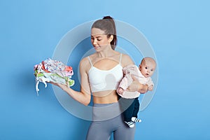 Cheerful European beautiful young woman holding baby girl in hands, carries stack of child`s clothing, looking at kid`s stuff,