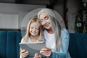 Cheerful european little girl and old woman watch video in tablet, read book online in living room interior