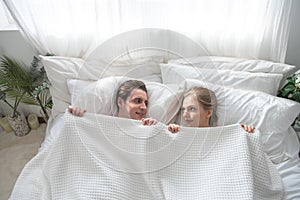 Cheerful excited Caucasian married couple lying in bed hiding bodies under white blanket