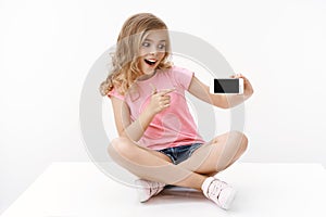 Cheerful excited cute blond little girl sitting on floor with crossed legs, hold smartphone, pointing mobile phone