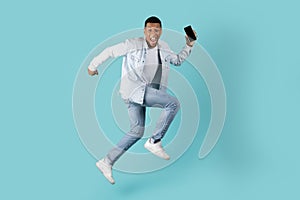 Cheerful excited millennial black guy with open mouth running, jumping with smartphone, has fun