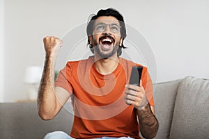 Cheerful excited young arabic male with open mouth with remote control make victory gesture and celebrates success