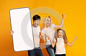 Cheerful Excited Young Family With Little Daughter Holding Big Blank Smartphone