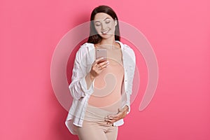 Cheerful expectant mother using mobile phone and chatting with her husband, touching her pregnant belly and smiling happily, cute