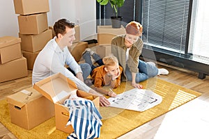 Cheerful family with child boy sitting on floor among cardboard boxes in new house, using blueprint