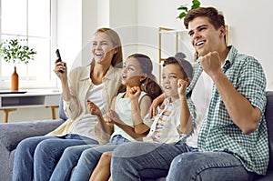 Cheerful family with children cheering together while watching TV at home in living room.