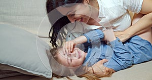 Cheerful family mother and daughter embracing and laughing lying on bed together. Happy young mom tickling her cute daughter