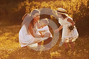 Cheerful family of mother, little son and daughter spending free time on the field at sunny day time of summer