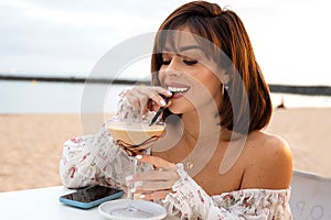 Cheerful fashionable brunette girl drinking ice coffee in beach bar, relaxing, using mobile phone and smiling