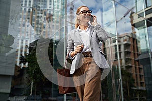 Cheerful fashionable business woman wearing eyeglasses and classic wear talking on mobile phone and smiling on the way