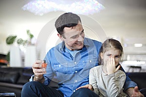 Cheerful father playing with little daughter sitting on couch and helping to paint her nails with bright polish play together