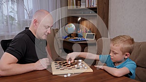 Cheerful father and son play checkers while sitting at a wooden table at home.