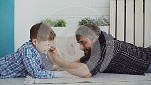 Cheerful father is teaching his little son arm wrestling, showing him hand position and pretenting to lose, boy is