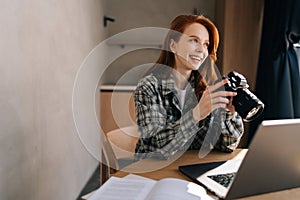 Cheerful female artist adjusting new professional photo camera or choosing photos for editing sitting in front of laptop