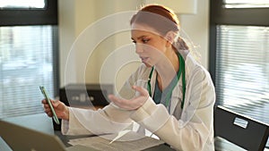 Cheerful female doctor having video call via mobile phone giving distant online consultation to patient and working with
