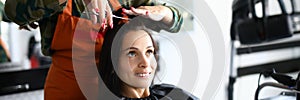 Cheerful female hairdresser in apron cut hair shortly to visitor with scissors.