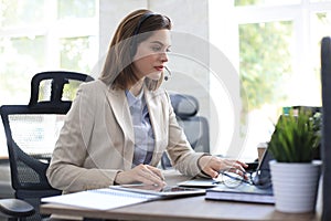 Cheerful female manager sitting at office desk and performing corporate tasks using wireless connection on gadgets