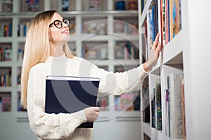 Cheerful female student with glasses standing near bookshelves in modern interior library of university.
