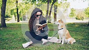 Cheerful female student is reading book in the park sitting on lawn and caressing beautiful dog, pet is yawning girl is