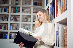 Cheerful female student standing near bookshelves in modern interior library of university during break between lesson.