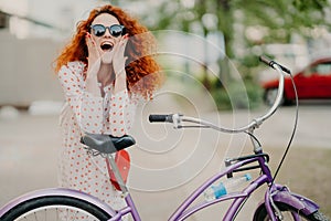 Cheerful female urban biker wears sunglasses and dress, has luxurious crisp foxy hair, keeps elbows on saddle of bicycle, spends