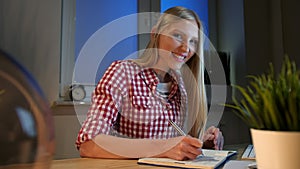 Cheerful female writing in daily planner. Lively young blond woman in casual checkered shirt sitting at wooden desk with