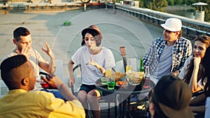 Cheerful friends are chatting and drinking lemonade sitting on rooftop on warm sunny day with snacks and drinks on table